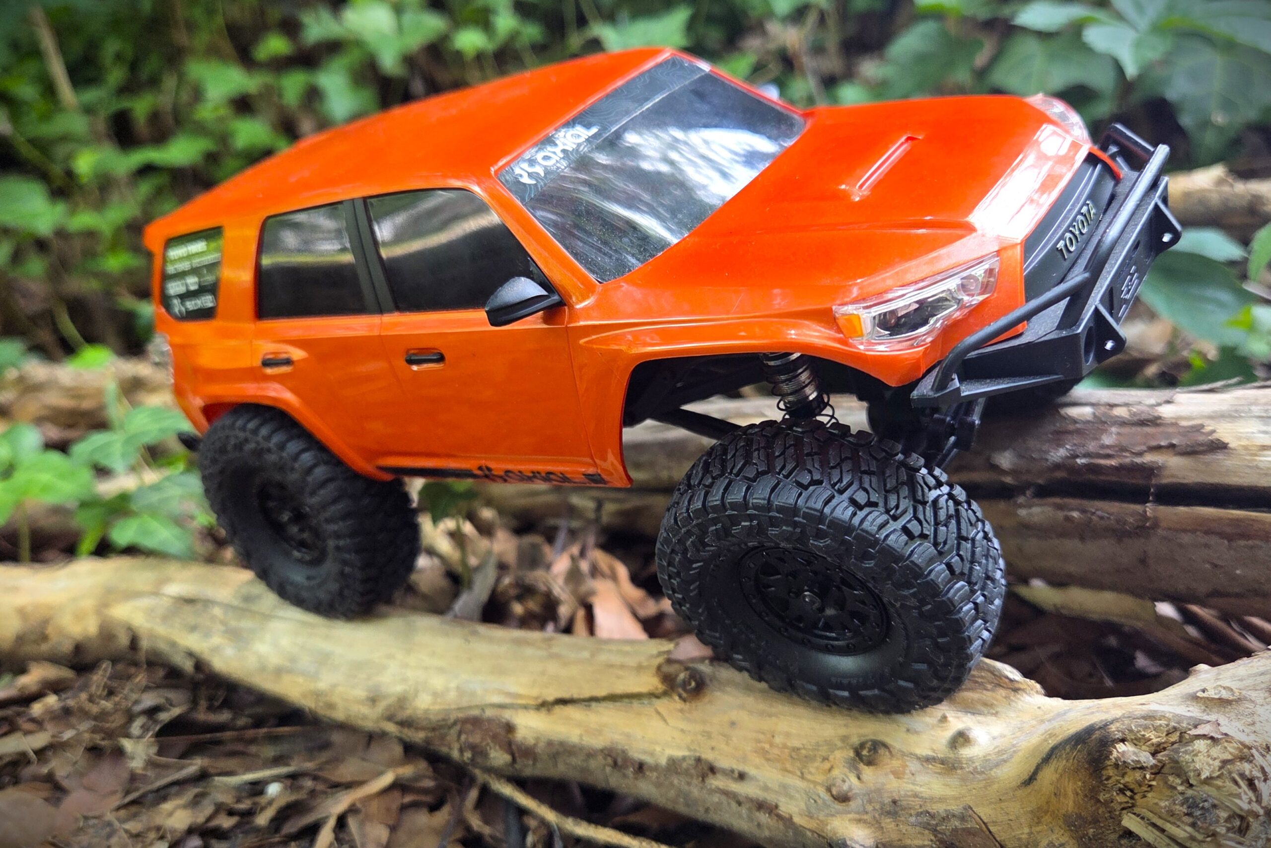 Orange Axial SCX24 Toyota 4Runner crawling over a log in the woods — suspension fully articulated