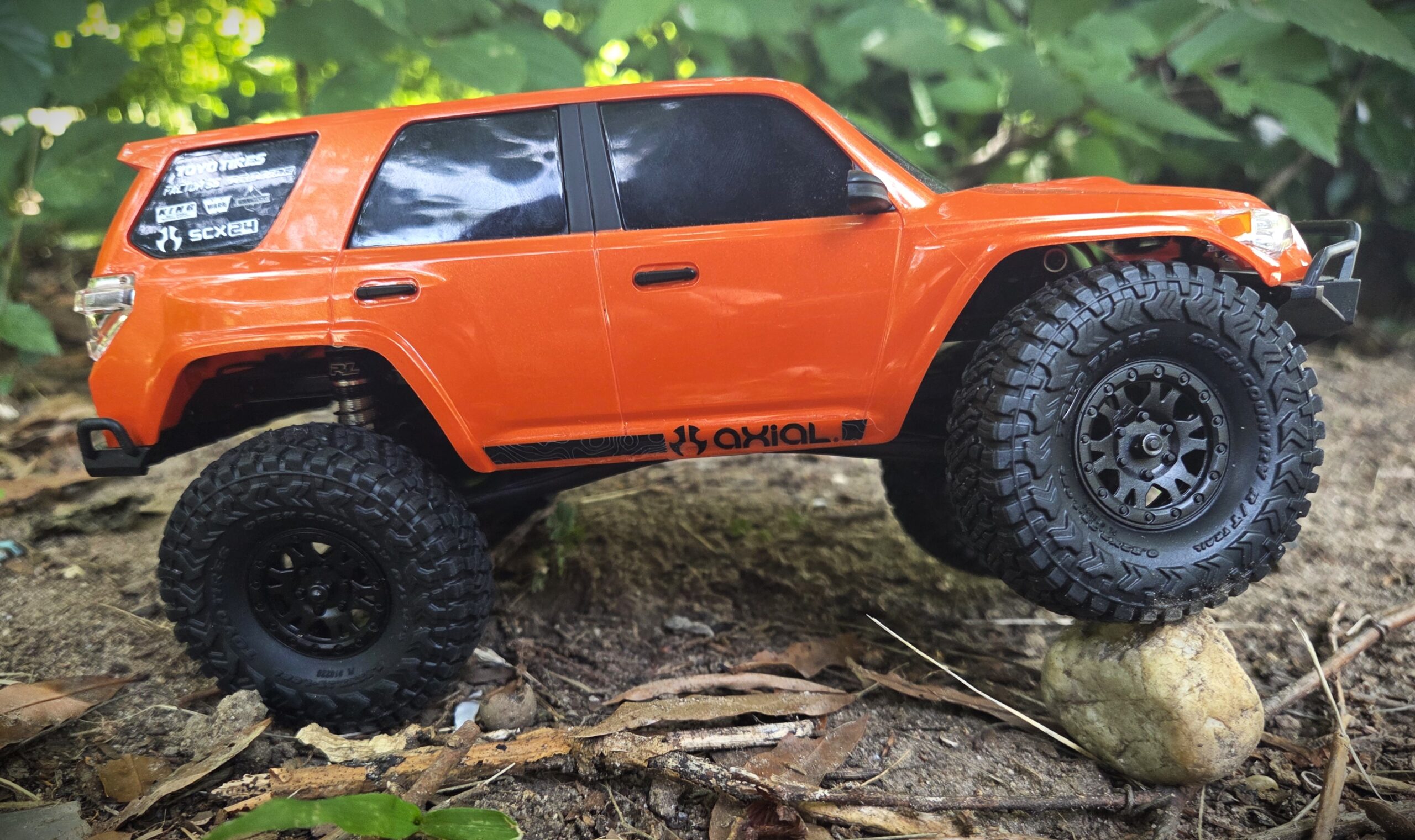 Orange Axial SCX24 Toyota 4Runner crawling over a rock in the woods, suspension fully articulated