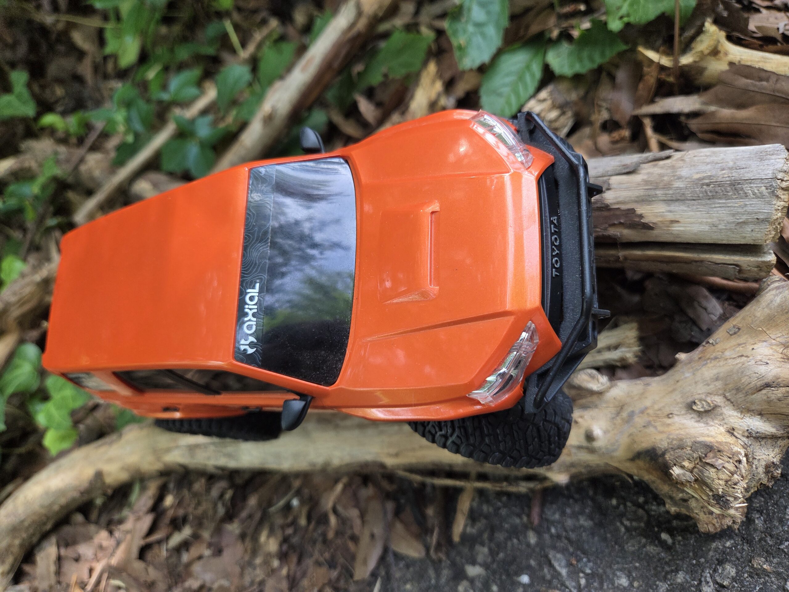 Orange Axial SCX24 Toyota 4Runner crawling over a large log in the woods, full side view