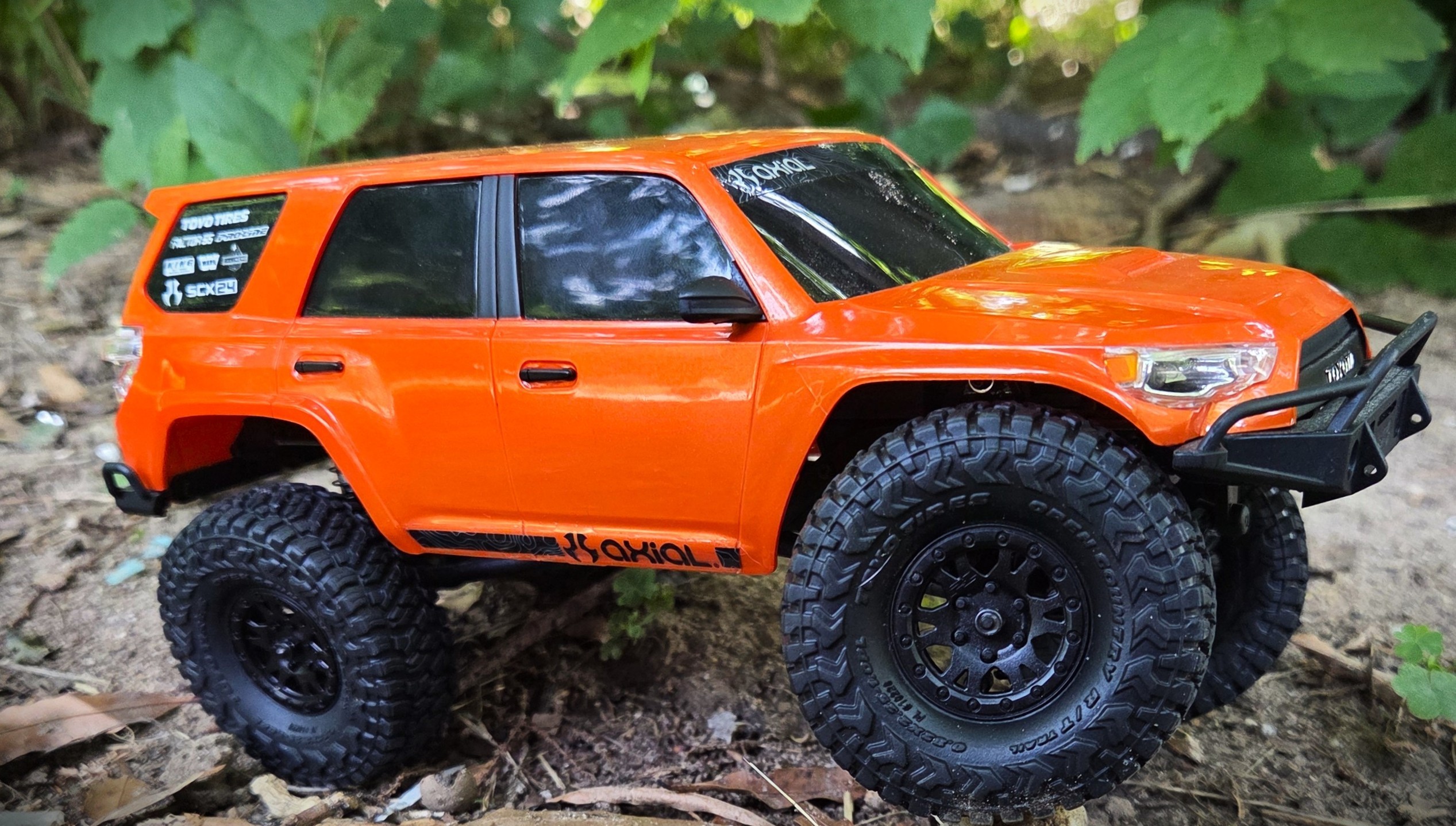 Axial SCX24 Toyota 4Runner in orange, side profile shot against green leaves backdrop